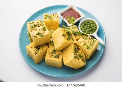 Soft and fluffy yellow Surti Khaman topped with mustard seeds, green chilies, coriander leaves, and grated coconut, served on a plate as a popular snack from Surat.” 😋